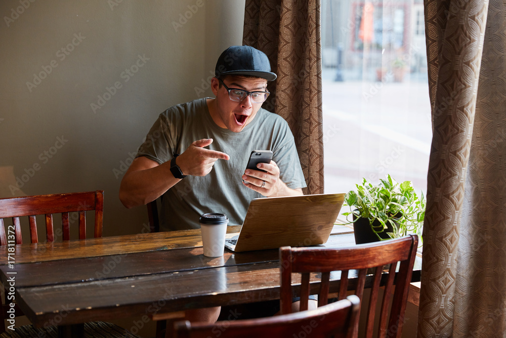 man on smart phone excited next to computer at coffee shop