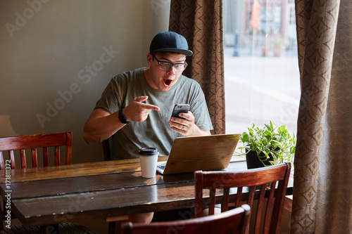 man on smart phone excited next to computer at coffee shop