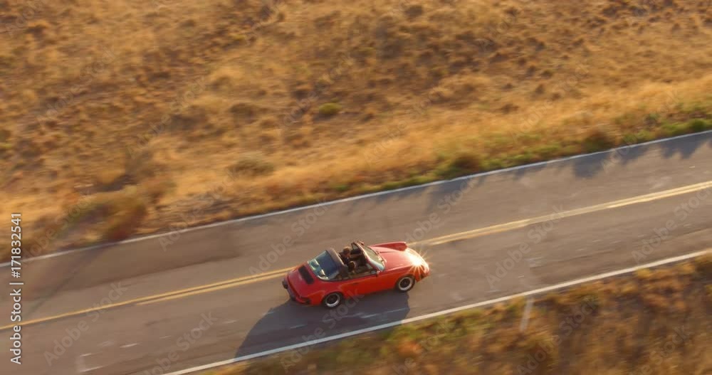 Aerial View Of Couple Driving Red Convertible Sports Car Down Country Road At Sunset 