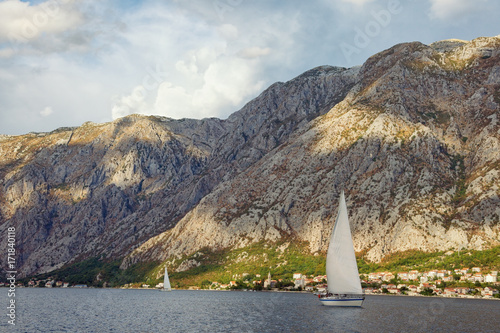 Fototapeta Naklejka Na Ścianę i Meble -  Sailboats sail along the shore of the Bay of Kotor near mountains covered with sunlight spots. Montenegro