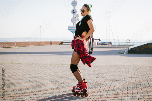 Young stylish funky girl with green hair riding roller skates and dancing near sea port during sunse