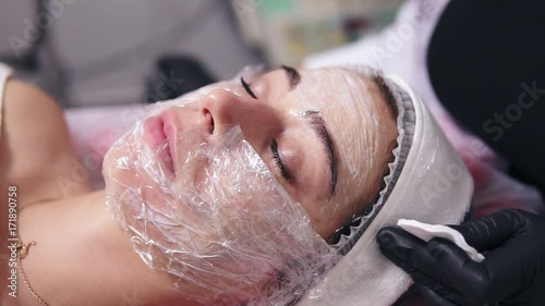 Professional cosmetologist is cleaning woman's face from special treatment using cotton sponge. Young woman is lying on the couch during cosmetic face procedure in spa salon photo