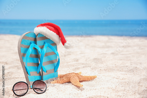 Fototapeta Naklejka Na Ścianę i Meble -  Santa hat with sea star, flip flops and sunglasses on beach. Christmas holidays concept
