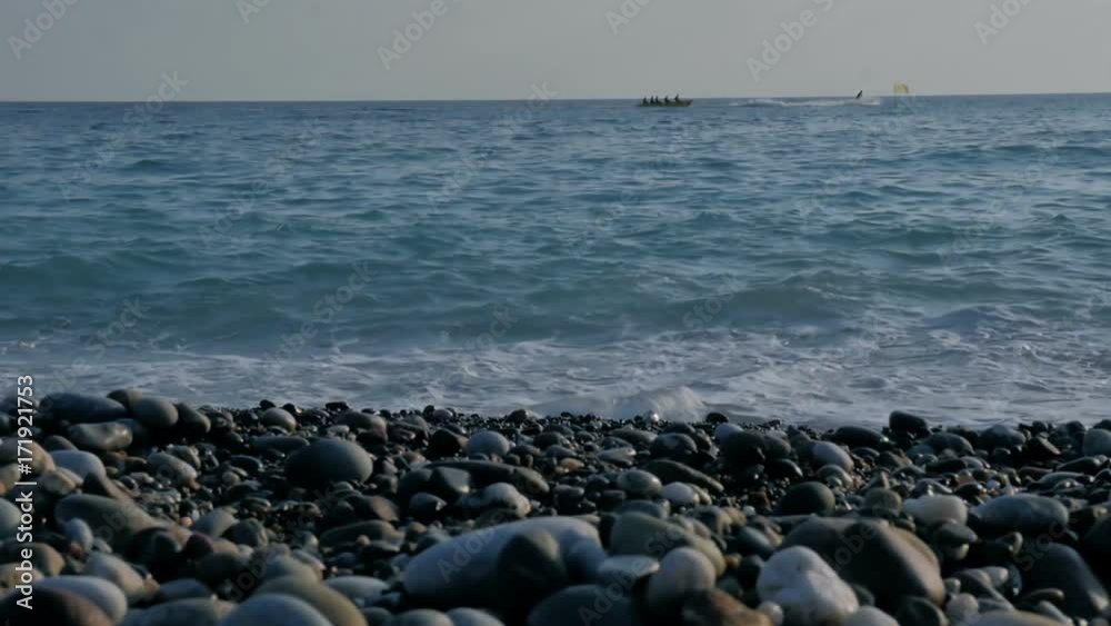 People ride on an inflatable banana by sea. Large transparent waves with foam. Sea pebble beach with colorful stones. People ride on an inflatable banana by sea. Large transparent waves with foam. Sea pebble beach with colorful stones.