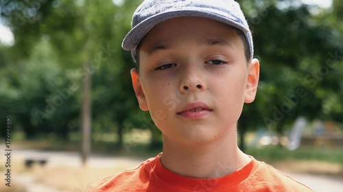 A kind seven-year-old boy with deep eyes and a cap on his head stands in a park on a picturesque lawn and thinks about his life with on a sunny day in summer in slow motion