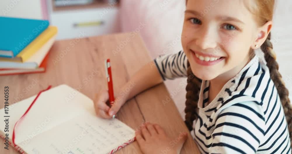 Portrait cheerful student girl doing homework. Child sitting at the table in the bedroom. Top view Portrait cheerful student girl doing homework. Child sitting at the table in the bedroom. Top view