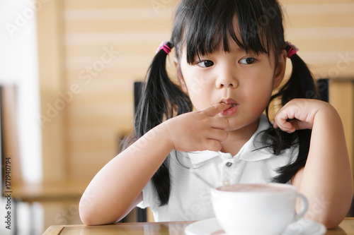 Asian children cute or kid girl nail biting with suck finger and delicious hot cocoa or chocolate drinking in white cup and looking for breakfast in the morning on table in home or cafe restaurant