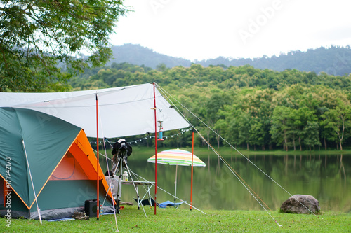 Fototapeta Naklejka Na Ścianę i Meble -  nature orange with green camping tent and gray fly sheet with tent accessory on meadow with lake or river and tree mountain for holiday relax on rainy season and white sky at ched khot