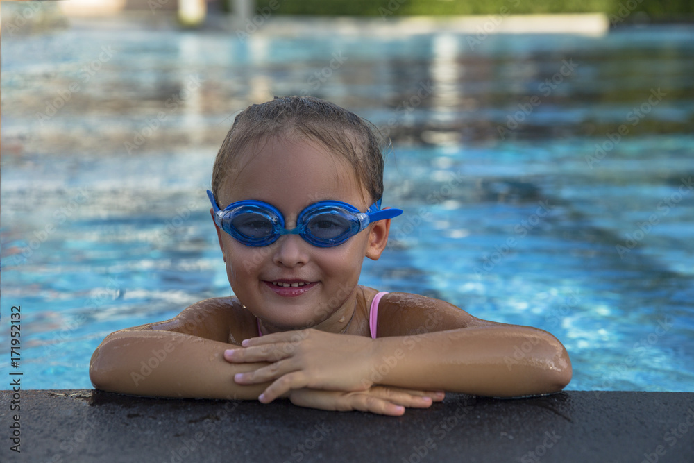 A happy young girl relaxing on the side of a swimming pool wearing goggles