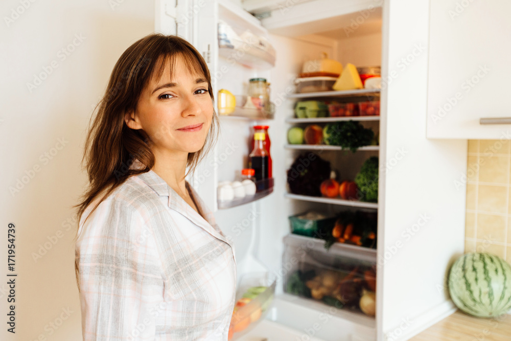 Young beautiful brunette woman in pajamas searching for food in the fridge and looking at camera.