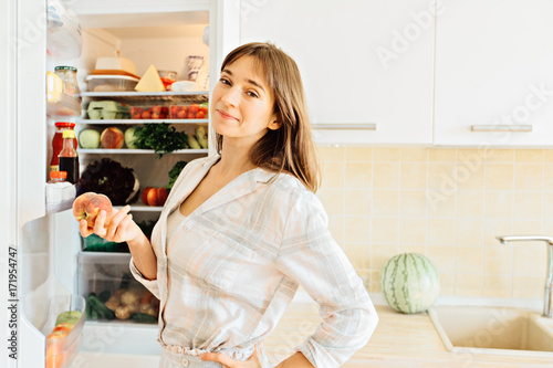 Cute female in pajamas taking peach from the fridge, attractive housewife take care about health, fresh tasty organic food, healthy eating concept.