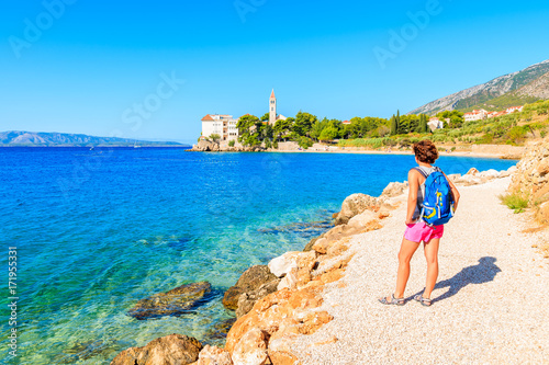 Young woman tourist standing on coastal path along sea and looking at distant Dominican monastery in Bol town, Brac island, Croatia