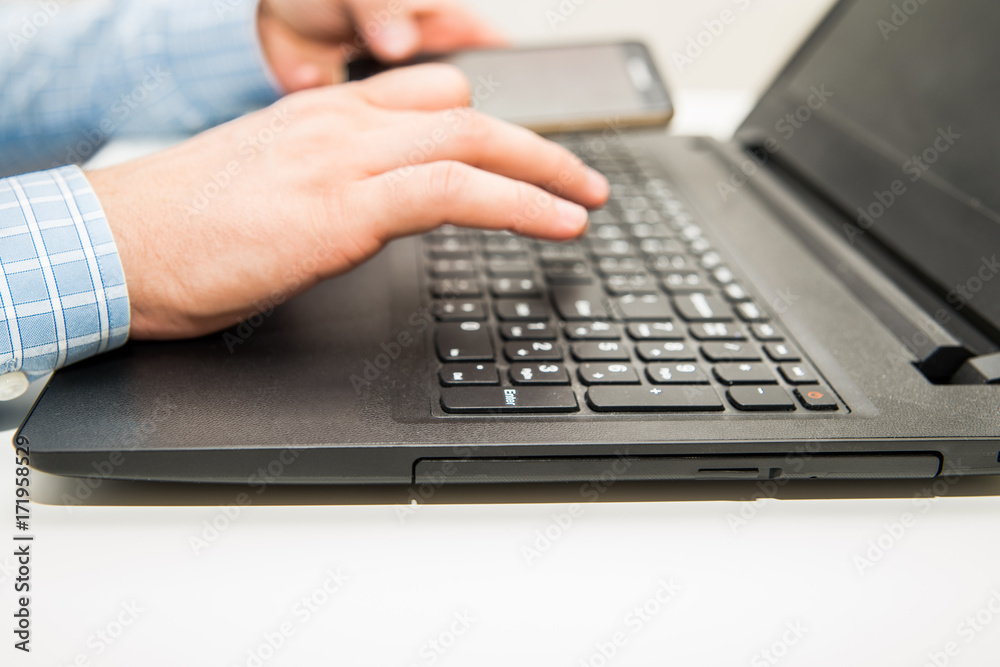 Man writes on keyboard holding mobile phone in other hand. Man sitting in front of a laptop and working on a computer in his office work. Man writes on keyboard holding mobile phone in other hand. Man sitting in front of a laptop and working on a computer in his office work.