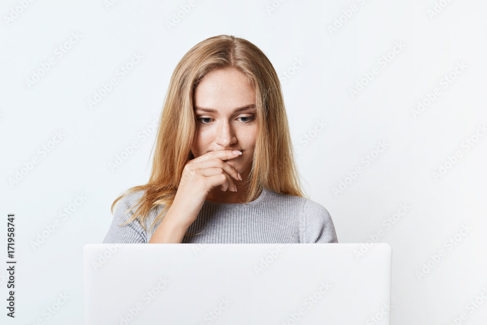 Puzzled female with beautiful appearance reads news online, being focused on laptop computer. Young college student works at his diploma paper or thesis, uses modern technology, isolated on white Puzzled female with beautiful appearance reads news online, being focused on laptop computer. Young college student works at his diploma paper or thesis, uses modern technology, isolated on white