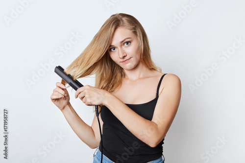 Fashionable woman does hairstyle with hair straightener, going to birthday party or has date with boyfriend, tries to look wonderful and appealing. Female combs perfect hair with iron. Hair care
