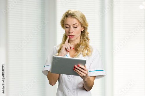 Thinking nurse using digital tablet. Beautiful young female doctor or nurse reading news on computer tablet and thinking.