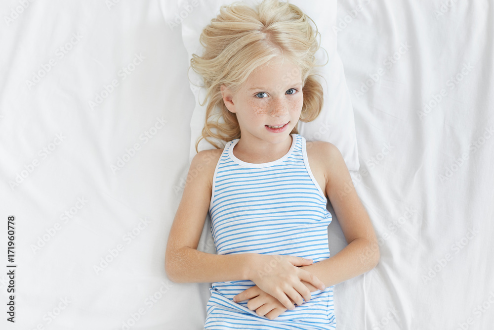 Top view of charming little blonde girl with cute freckles all over her face lying on pillow in her white bed looking and smiling at camera, clasping hands in front of her. Childhood and happiness