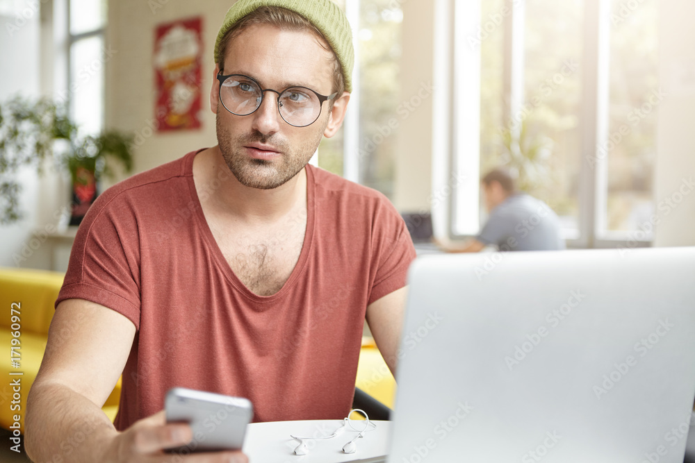 Fashionable man texts sms on smart phone to girlfriend, waits her in cafeteria or coffee shop, enjoys free internet connection, sits in front of generic laptop. Male freelancer works remotely Fashionable man texts sms on smart phone to girlfriend, waits her in cafeteria or coffee shop, enjoys free internet connection, sits in front of generic laptop. Male freelancer works remotely