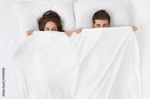 Obraz na plátně Funny married couple lying in bed and hiding under white blanket, looking at camera with eyes full of joy