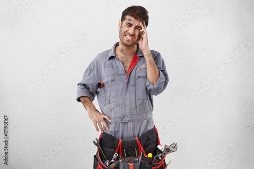 Handsome young bearded Caucasian mechanic wearing blue work clothes touching head, having painful look, feeling exhausted or stressed after hard working day. Negative human facial expressions