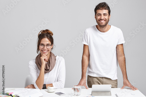 Two successful creative colleagues working together at white office desk. Cheerful unshaven male architect duscussing new architectural project with his female colleague, gadgets and papers on table