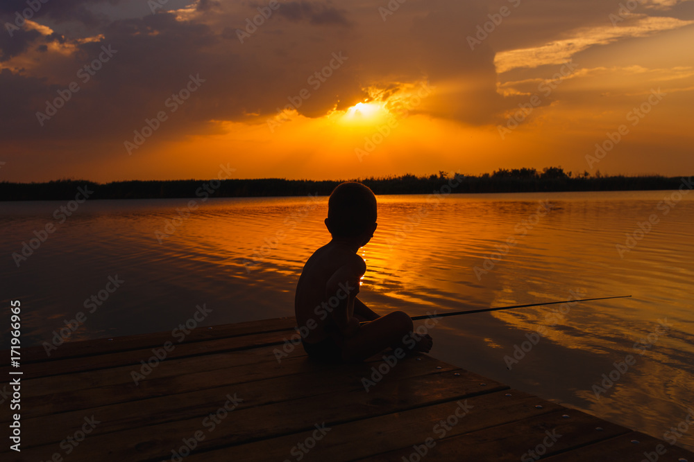 Little boy siting on wooden dock and fishing at sunset.