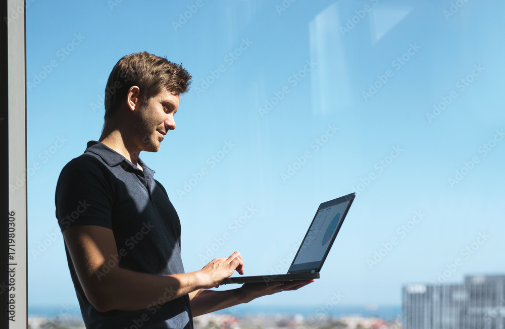 Smiling young casual businessman holding and using laptop with blurred city background