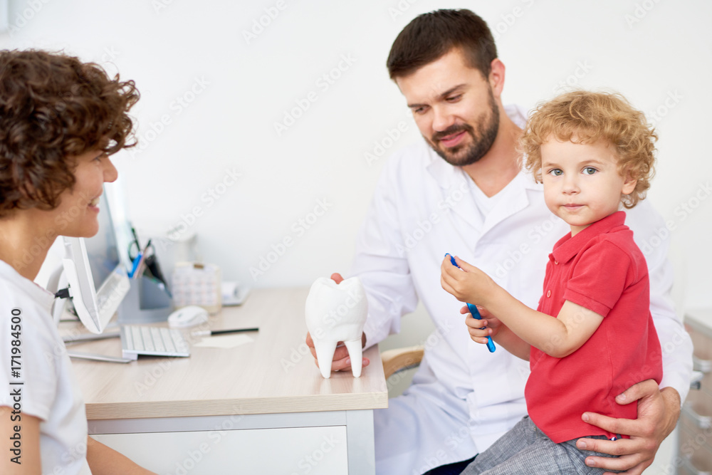 Portrait of adorable little boy looking at camera while sitting on laps of handsome bearded dentist and learning to brush teeth correctly Portrait of adorable little boy looking at camera while sitting on laps of handsome bearded dentist and learning to brush teeth correctly