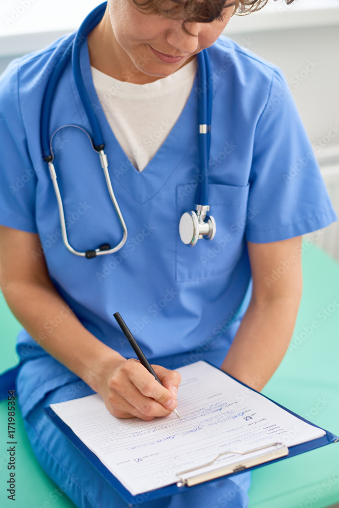 Confident middle-aged doctor wearing blue uniform filling in medical record while sitting at modern office, close-up shot Confident middle-aged doctor wearing blue uniform filling in medical record while sitting at modern office, close-up shot