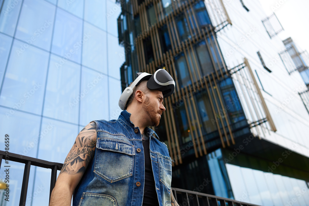 People, science, gaming, simulation and augmented reality. Serious confident young man with stubble and stylish tattoos posing in urban surroundings, using oculus rift headset for entertaining himself People, science, gaming, simulation and augmented reality. Serious confident young man with stubble and stylish tattoos posing in urban surroundings, using oculus rift headset for entertaining himself
