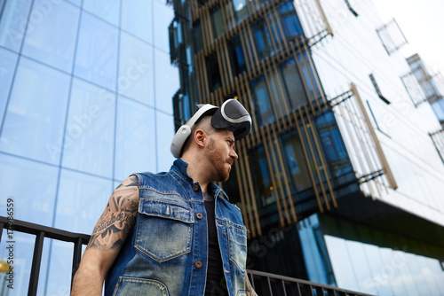 People, science, gaming, simulation and augmented reality. Serious confident young man with stubble and stylish tattoos posing in urban surroundings, using oculus rift headset for entertaining himself