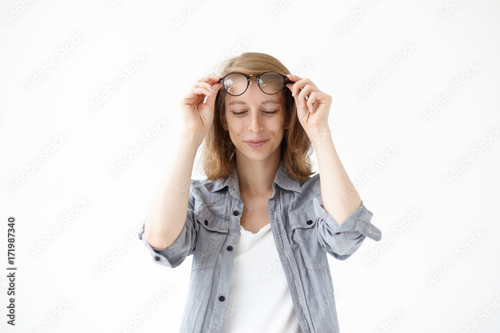 Charming young female in casual wear holding her big spectacles in hands and looking down with shy cute smile. Pretty girl with freckles posing in studio, putting off eyeglasses in black frame Charming young female in casual wear holding her big spectacles in hands and looking down with shy cute smile. Pretty girl with freckles posing in studio, putting off eyeglasses in black frame