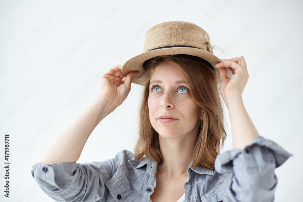 People, clothing, design, style, fashion and shopping concept. Isolated studio picture of gorgeous blue-eyed young woman with freckles trying on vintage round hat while shopping at flea market People, clothing, design, style, fashion and shopping concept. Isolated studio picture of gorgeous blue-eyed young woman with freckles trying on vintage round hat while shopping at flea market