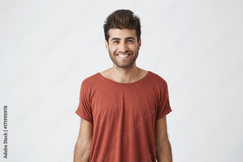 Young cheerful hispanic guy in red t-shirt brightly smiling hearing good news from friend. Beardy handsome student looking at camera with joyful smile Young cheerful hispanic guy in red t-shirt brightly smiling hearing good news from friend. Beardy handsome student looking at camera with joyful smile