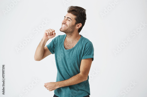 Indoor portrait of expressive spanish mature guy with stylish haircut in blue t-shirt, brightly smiling, singing, opening her mouth widely and clothing eyes. Body language