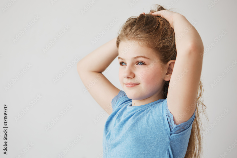 Beautiful tiny girl with charming blue eyes and long light hair smiles, looking aside and making hairstyle. Childhood, girlhood and lifestyle concept.