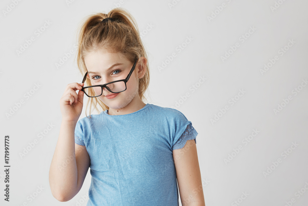 Cute small blonde girl with blue eyes and pleasant smile in blue t-shirt funny posing with new glasses for school photo.