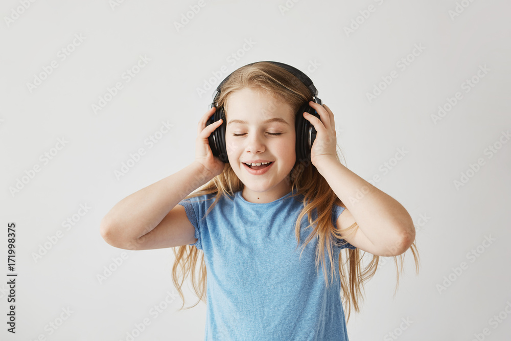 Portrait of little blonde girl in blue shirt playing with big wireless headphones, listening to music, singing song and dancing with closed eyes while nobody home. Portrait of little blonde girl in blue shirt playing with big wireless headphones, listening to music, singing song and dancing with closed eyes while nobody home.
