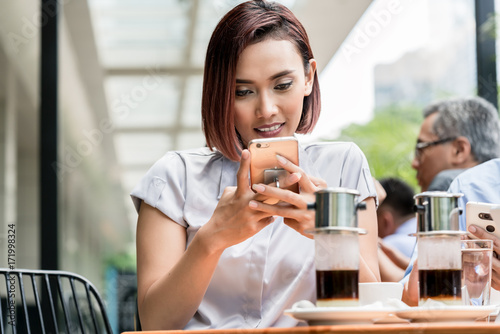 Portrait of a young Asian woman smiling while using a mobile phone connected to the internet outdoors at a coffee shop