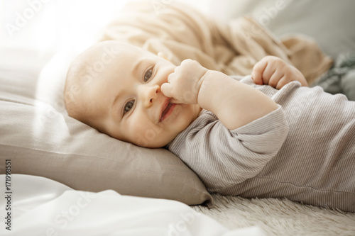 Portrait of sweet smiling newborn daughter lying on cozy bed. Child looks at camera and touching face with her little hands. Childhood moments.
