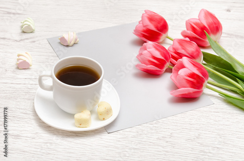 Pink tulips on a blank sheet of paper, mug of coffee and marshmallows, light wooden background