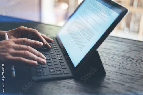 Closeup view of male hands typing on electronic tablet keyboard-dock station. Business text information on device screen. Man working at office.Horizontal side-view.