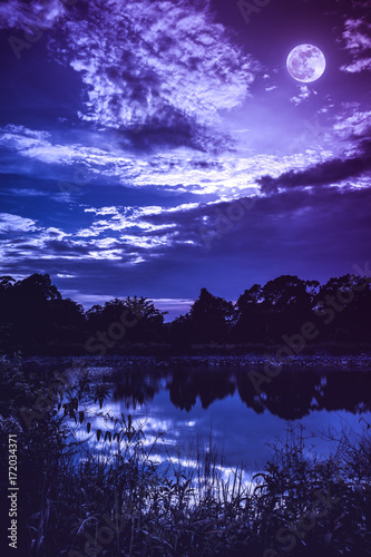 Sky with dark cloudy and full moon above silhouettes of trees and tranquil lake. © kdshutterman