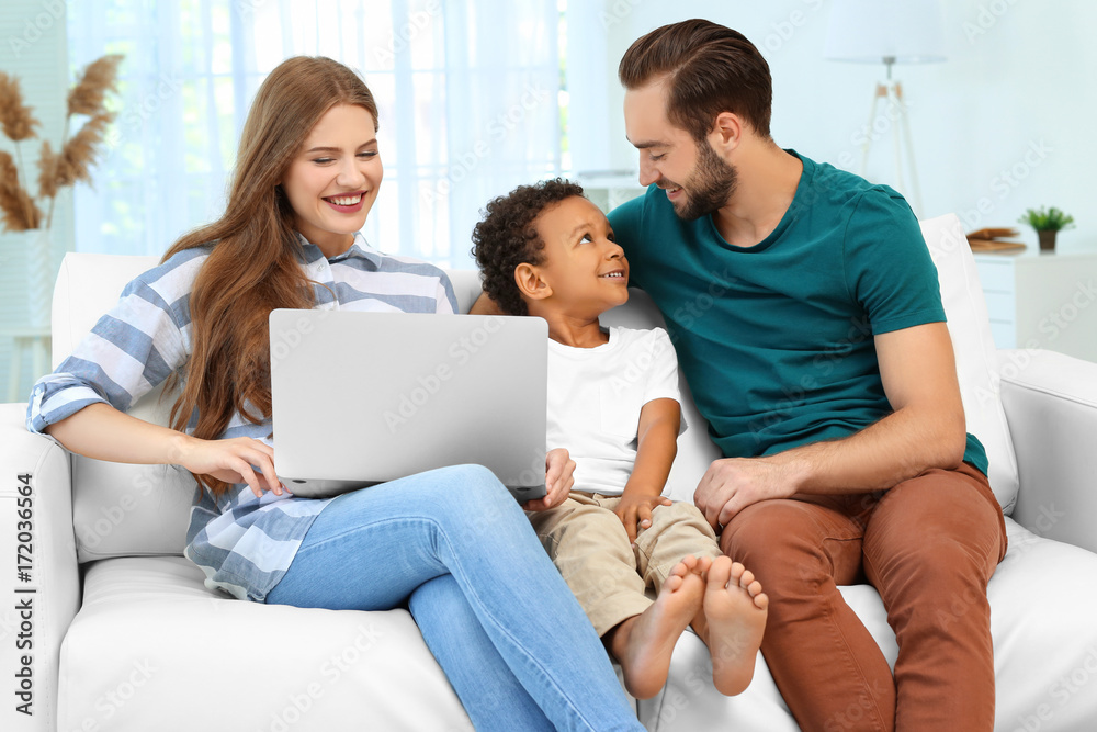 Happy couple with adopted African-American boy using laptop while sitting on couch at home