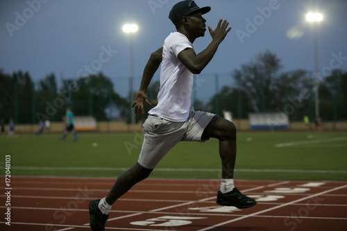 African male sportsman in a cap fast running short distance on stadium track, he trains speed © antgor