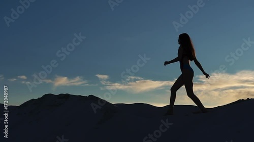 Woman in blue swimming suit walks on the sand dune in the evening