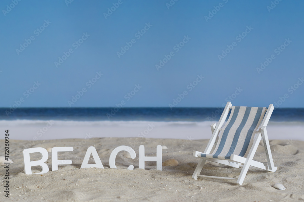 The word beach in white wooden letters in the sand on the beach, beside a deck chair