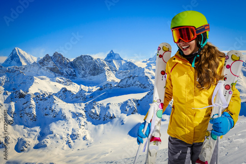 Skier teenager along a snowy ridge with skis. In background blue sky and shiny sun and Swiss Alps.  Adventure winter sport.