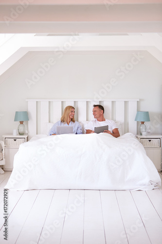 Couple Wearing Pajamas Lying In Bed Using Digital Devices