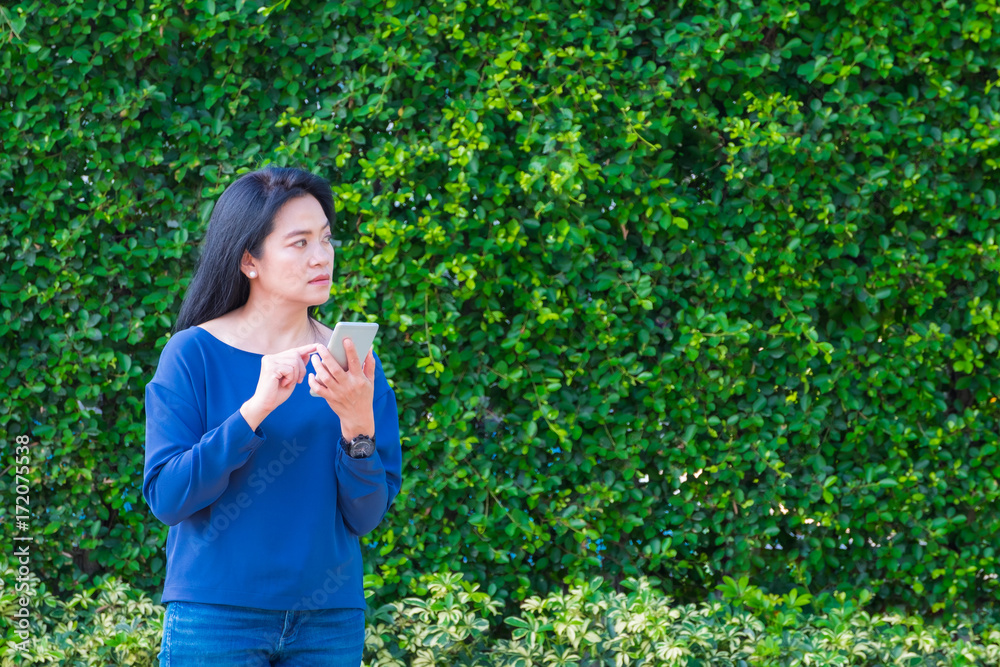 Asian woman in causal cloth and smile while use mobile phone outside house at green hedge,Leave copy space for adding your text.